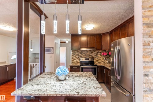 Kitchen featuring an island with a light-colored stone countertop, stainless steel appliances, dark wood cabinetry, a mosaic tile backsplash, and overhead pendant lighting - 16524 116 Street, Edmonton, AB 