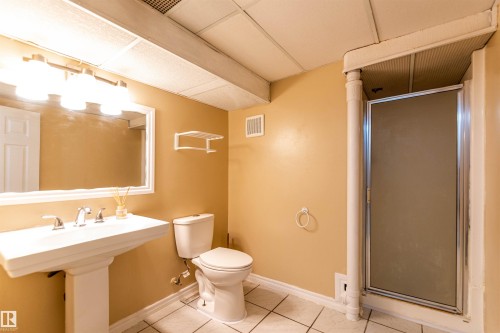 Bathroom featuring a pedestal sink with chrome fixtures, a mirrored vanity, a toilet, a shower stall with a frosted glass door, and tiled flooring - 16524 116 Street, Edmonton, AB 