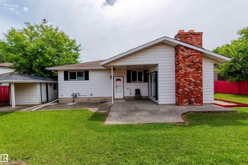 Rear exterior featuring white horizontal siding, a red brick chimney, a concrete patio, and a grass lawn - 16524 116 Street, Edmonton, AB 