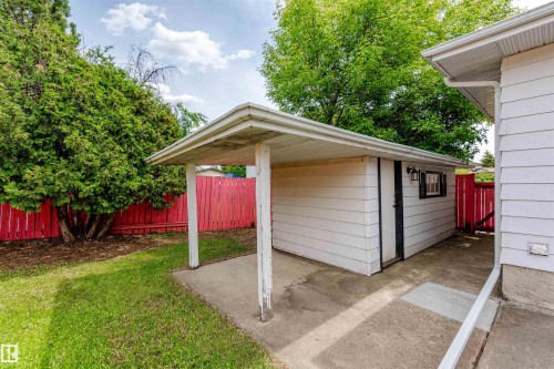 Detached storage shed with covered concrete patio, black framed window with shutters, and exterior lighting, alongside a red picket fence and mature landscaping - 16524 116 Street, Edmonton, AB 