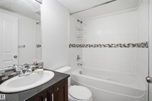 Bathroom featuring a white bathtub, white tiled shower surround with mosaic accent band, dark wood-finish vanity with an oval sink, and a large wall-mounted mirror - 115 274 Mcconachie Drive, Edmonton, AB 