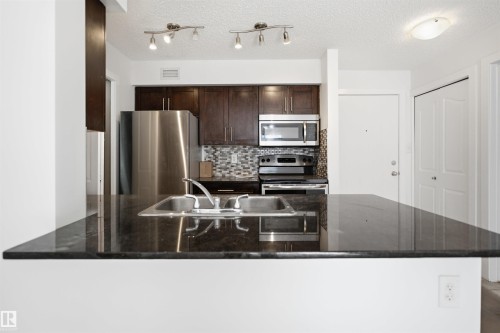 Kitchen featuring a double basin stainless steel sink, dark wood cabinetry, stainless steel appliances, and a mosaic tile backsplash - 115 274 Mcconachie Drive, Edmonton, AB 