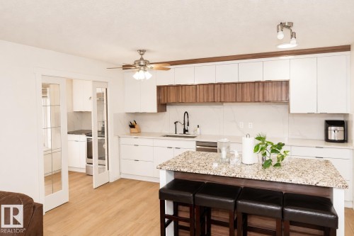 Contemporary kitchen featuring an island with a speckled countertop, white cabinetry with wood-finish accents, and light wood-finish flooring - 8523 7 Avenue Sw, Edmonton, AB 