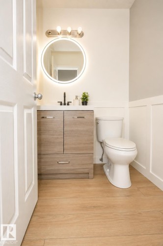 Powder room featuring a wood-finish vanity with a white countertop and a circular backlit mirror - 8523 7 Avenue Sw, Edmonton, AB 
