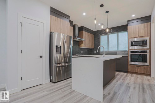 Kitchen featuring a waterfall-edge island with a gooseneck faucet, stainless steel appliances, dark brick-patterned tile backsplash, and light wood-finish flooring - 10628 81 Street Nw, Edmonton, AB 