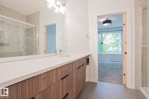 Spacious vanity area featuring a dual sink configuration with polished chrome faucets, a broad mirror, and a contemporary wood-finish cabinet with dark hardware - 10628 81 Street Nw, Edmonton, AB 