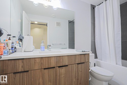 Bathroom featuring a wood-finish vanity with integrated sink, a large wall-mounted mirror, overhead vanity lighting, and a white bathtub with dark gray tile surround - 10628 81 Street Nw, Edmonton, AB 