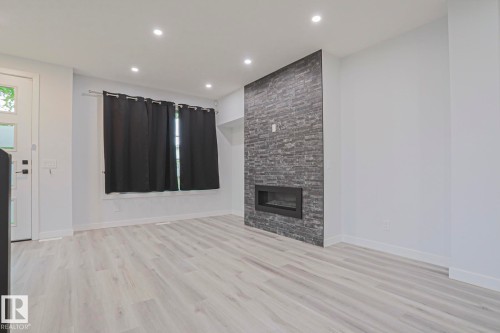 Living space featuring light wood-finish flooring, recessed ceiling lighting, a modern electric fireplace with a stacked stone-look surround, and a white entry door with frosted glass inserts - 10628 81 Street Nw, Edmonton, AB 