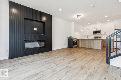 Open-concept living space with wood-finish flooring, a contemporary fireplace feature wall, a kitchen island with seating, stainless steel appliances, and white cabinetry - 11336 130 Street Nw, Edmonton, AB 