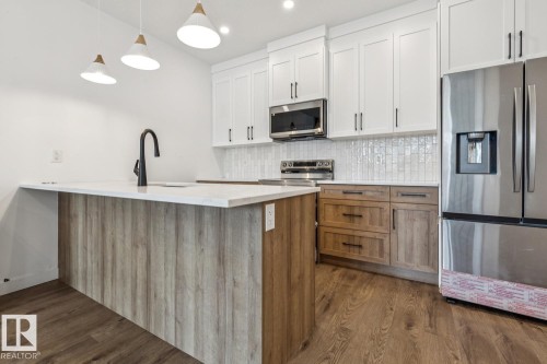 Kitchen featuring stainless steel appliances, white cabinetry, wood-finish lower cabinetry, a white countertop island with a matte black faucet, and wood-finish flooring - 11336 130 Street Nw, Edmonton, AB 