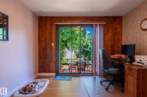 Interior space featuring wood-finish flooring, a full wall of vertical wood paneling, and a glass sliding door providing access to an outdoor patio with a dining set and surrounding greenery - 14708 108 Av, Edmonton, AB 