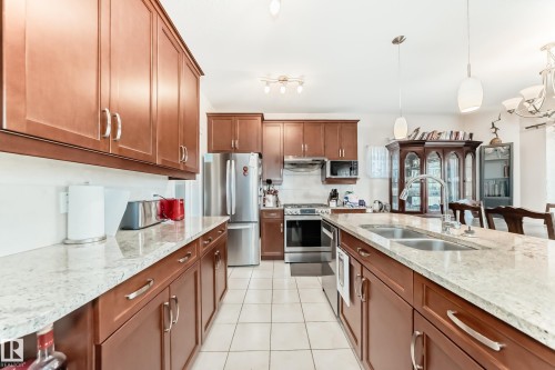 Kitchen featuring abundant wood cabinetry, light-toned countertops, stainless steel appliances, a center island with double sink, and tile flooring - 8730 180A Avenue, Edmonton, AB 