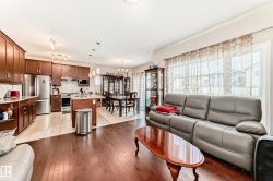 Open-concept main living area featuring wood-finish flooring, a kitchen with dark wood cabinetry and stainless steel appliances, a dining space with tiled flooring, and large windows with sheer coverings - 