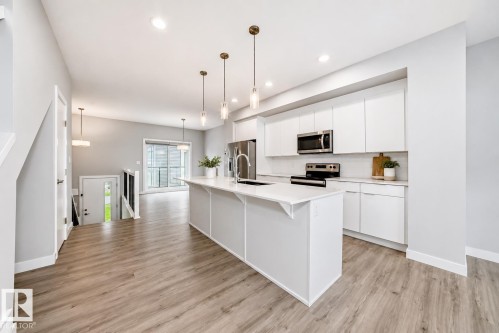 Modern kitchen featuring a white island with sink and three pendant lights, stainless steel appliances, white cabinetry, light countertops, and wood-finish flooring - 26 1729 Keene Crescent, Edmonton, AB 