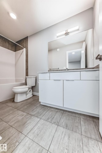 Spacious bathroom featuring a white vanity with multiple cabinet doors and drawers, a large wall-mounted mirror, overhead linear lighting, a white tub with tile surround, and wood-finish tile flooring - 26 1729 Keene Crescent, Edmonton, AB 