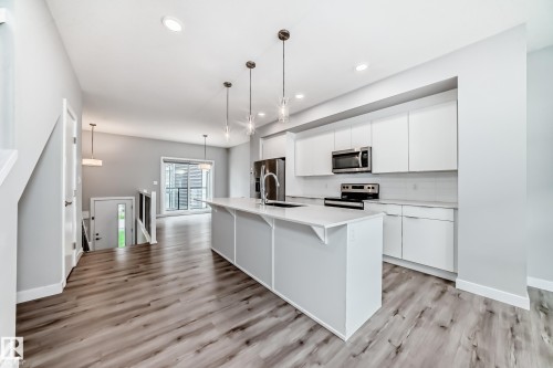 Modern kitchen featuring an expansive center island with pendant lighting, white cabinetry, stainless steel appliances, and wood-finish flooring - 26 1729 Keene Crescent, Edmonton, AB 