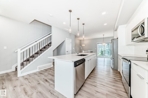 Open-concept kitchen featuring an elongated island with integrated sink, stainless steel appliances, white cabinetry, and wood-finish flooring - 26 1729 Keene Crescent, Edmonton, AB 
