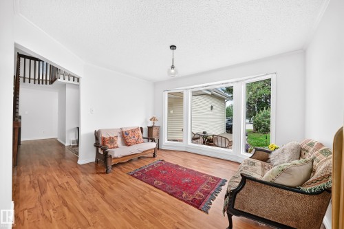 Bright living area featuring a large bay window, wood-finish flooring, a contemporary pendant light fixture, white walls, and a visible staircase with wood railings - 8224 186 Street Nw, Edmonton, AB 