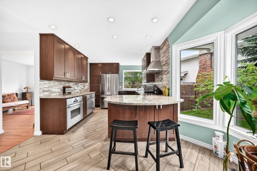 Modern kitchen featuring wood-finish cabinetry, stainless steel appliances, a tile backsplash, and a central island with a stone countertop - 8224 186 Street Nw, Edmonton, AB 