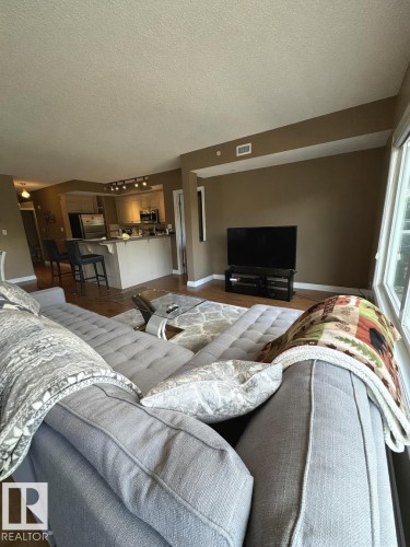 Open-concept living area featuring wood-finish flooring, a kitchen with track lighting, an island with seating, and multiple windows providing natural light - 305 11111 82 Avenue, Edmonton, AB 
