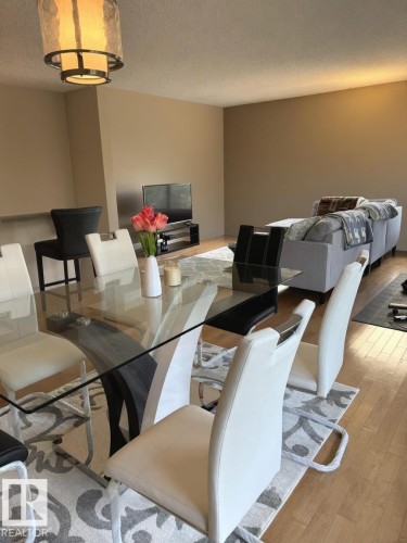 Open-concept living space featuring wood-finish flooring, a contemporary glass-top dining table, and a tiered drum chandelier - 305 11111 82 Avenue, Edmonton, AB 