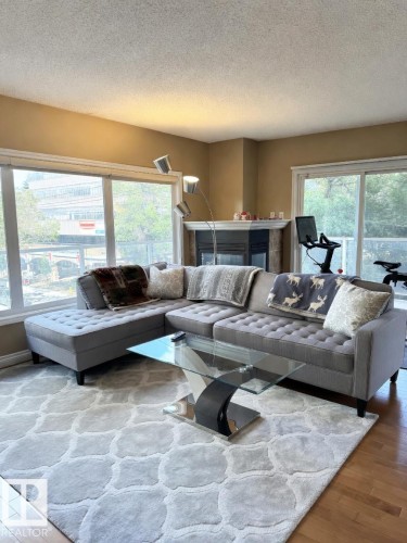 Living area featuring wood-finish flooring, a corner fireplace with a dark-toned surround and white mantel, and large windows with white trim - 305 11111 82 Avenue, Edmonton, AB 