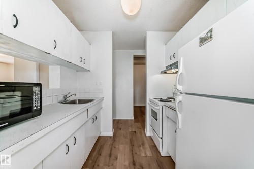 Galley kitchen featuring white cabinetry, white appliances including a refrigerator and electric range, laminate countertops, a stainless steel sink, and wood-finish flooring - 401 11007 83 Avenue, Edmonton, AB 