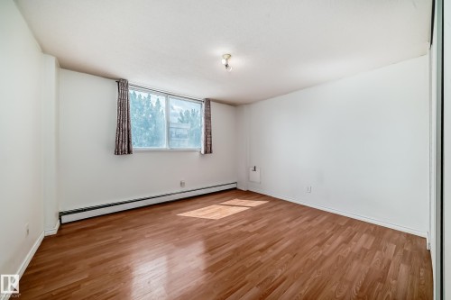 Spacious room featuring wood-finish flooring, a large window, white walls, baseboard heating, and a built-in closet with mirrored doors - 401 11007 83 Avenue, Edmonton, AB 