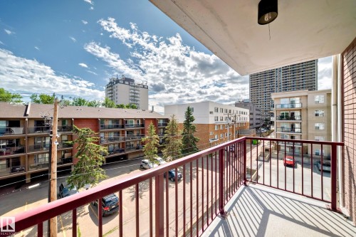 Private balcony with a red metal railing, offering urban surroundings with various building styles and mature trees - 401 11007 83 Avenue, Edmonton, AB 
