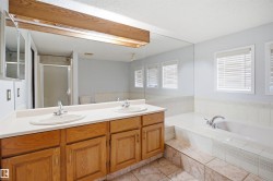Bathroom featuring a dual vanity with wood cabinetry, a large built-in mirror, a tiled soaking tub, and multiple windows with blinds - 