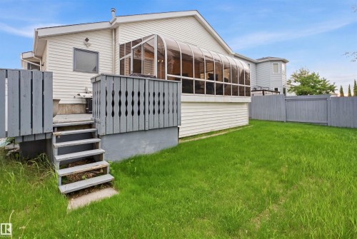 Rear exterior featuring a sunroom addition with curved glass panels, white siding, a multi-level deck with gray railings, steps leading to a grassy yard, and a gray privacy fence - 3220 42A Avenue, Edmonton, AB 