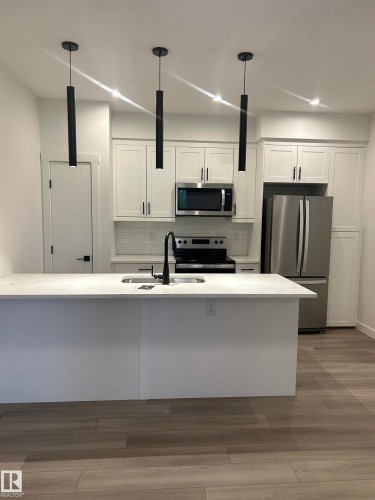 Modern kitchen featuring a large island with a light-toned countertop, white cabinetry with dark hardware, a stainless steel appliance suite, subway tile backsplash, and wood-finish flooring - 11012 132 Street, Edmonton, AB 