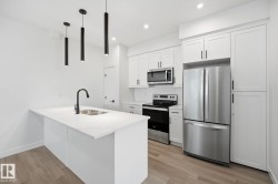 Kitchen featuring a white island with an undermount double sink and matte black faucet, pendant lighting, wood-finish flooring, stainless steel appliances, and white shaker cabinetry - 