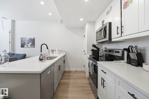 Contemporary kitchen featuring a peninsula with a stainless steel sink and matte black faucet, white shaker cabinetry with black hardware, light wood-finish flooring, a white subway tile backsplash, and stainless steel appliances - 11007 132 Street, Edmonton, AB 