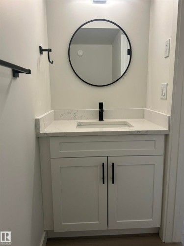 Bathroom vanity featuring white shaker-style cabinetry, a stone countertop with an integrated sink, a matte black faucet, and a circular mirror with a black frame - 11007 132 Street, Edmonton, AB 