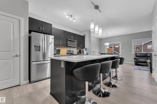 Open-concept kitchen featuring an island with seating, dark wood-finish cabinetry, stainless steel appliances, and light wood-finish flooring - 9328 Pear Link Link, Edmonton, AB 