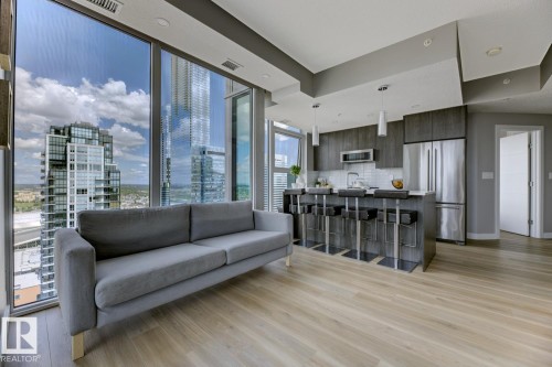 Open-concept living space featuring expansive floor-to-ceiling windows, light wood-finish flooring, a contemporary kitchen with dark cabinetry, stainless steel appliances, and a central island with seating - 2902 10180 103 Street, Edmonton, AB 