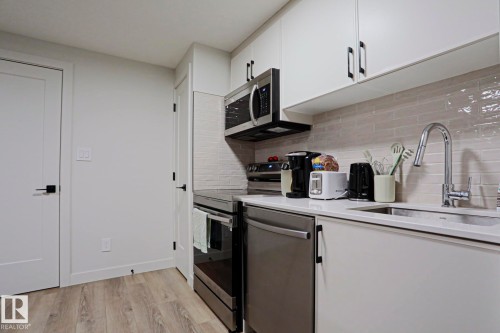 Modern kitchen featuring stainless steel appliances, white flat-panel cabinetry, a light-toned horizontal tile backsplash, wood-finish flooring, and a chrome gooseneck faucet - 12842 121 Street, Edmonton, AB 
