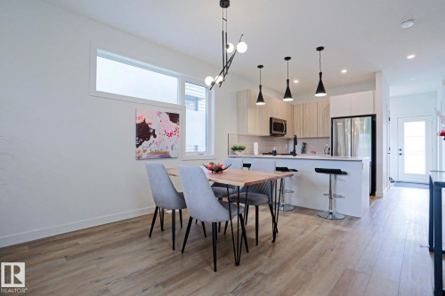 Open-concept living space featuring wood-finish flooring, a modern kitchen island with bar seating, stainless steel appliances, and a sleek linear chandelier - 12842 121 Street, Edmonton, AB 