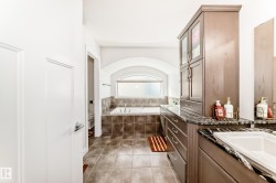 Bathroom featuring a built-in soaking tub with tile surround, arched window, and dual vanity with dark cabinetry and stone countertops - 