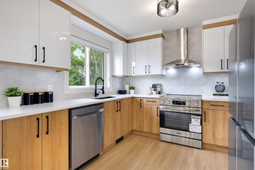 Modern kitchen featuring two-tone cabinetry with white upper cabinets and natural wood-finish lower cabinets, stainless steel appliances, white countertops, a hexagon-tiled backsplash, and wood-finish flooring - 12611 47 Street, Edmonton, AB 