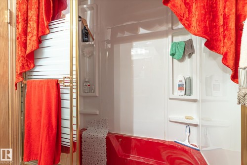 Red bathtub with integrated white shower walls featuring built-in shelving, complemented by a gold-framed shower door - 9747 67 Avenue, Edmonton, AB 