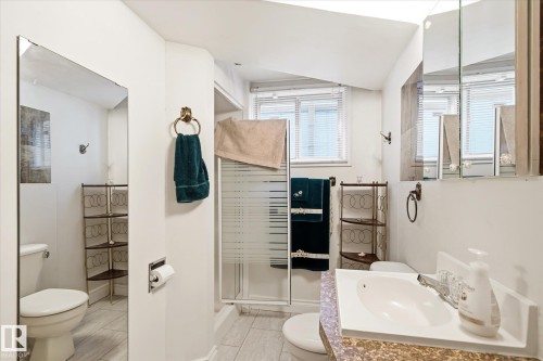 Bathroom featuring a shower enclosure with a frosted panel, single vanity with a white basin and patterned countertop, and a window with horizontal blinds - 9747 67 Avenue, Edmonton, AB 