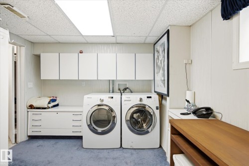Dedicated utility space featuring a front-loading washer and dryer, extensive white cabinetry, a built-in folding counter, a drop ceiling with recessed lighting, and soft blue carpeting - 9747 67 Avenue, Edmonton, AB 