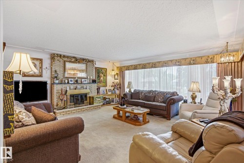 Spacious living area featuring a brick fireplace, light-toned carpeting, and a large window with sheer and fabric drapery - 9747 67 Avenue, Edmonton, AB 