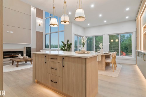 Open-concept living space featuring a central island with a light wood-finish base and light-toned countertop, a multi-pane window wall, recessed ceiling lighting, a contemporary fireplace, and wood-finish flooring - 3305 Chickadee Drive, Edmonton, AB 