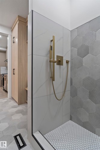 Walk-in shower featuring a brass-finish shower system, large format white tile with subtle veining, and a complementary hexagonal textured tile accent wall - 3305 Chickadee Drive, Edmonton, AB 
