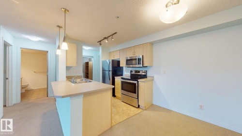 Open-concept kitchen featuring light wood-finish cabinetry, stainless steel appliances, and an island with a double basin sink and pendant lighting - 209 13908 136, Edmonton, AB 