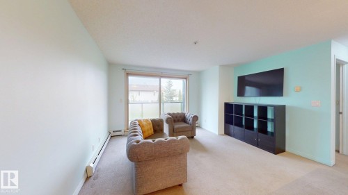 Spacious living area featuring carpet flooring, a light-colored accent wall, and a sliding glass door providing natural light - 209 13908 136, Edmonton, AB 