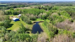 Expansive acreage featuring a private pond, mature trees, a main residence with an A-frame roof, and an additional blue outbuilding, all surrounded by natural landscaping - 